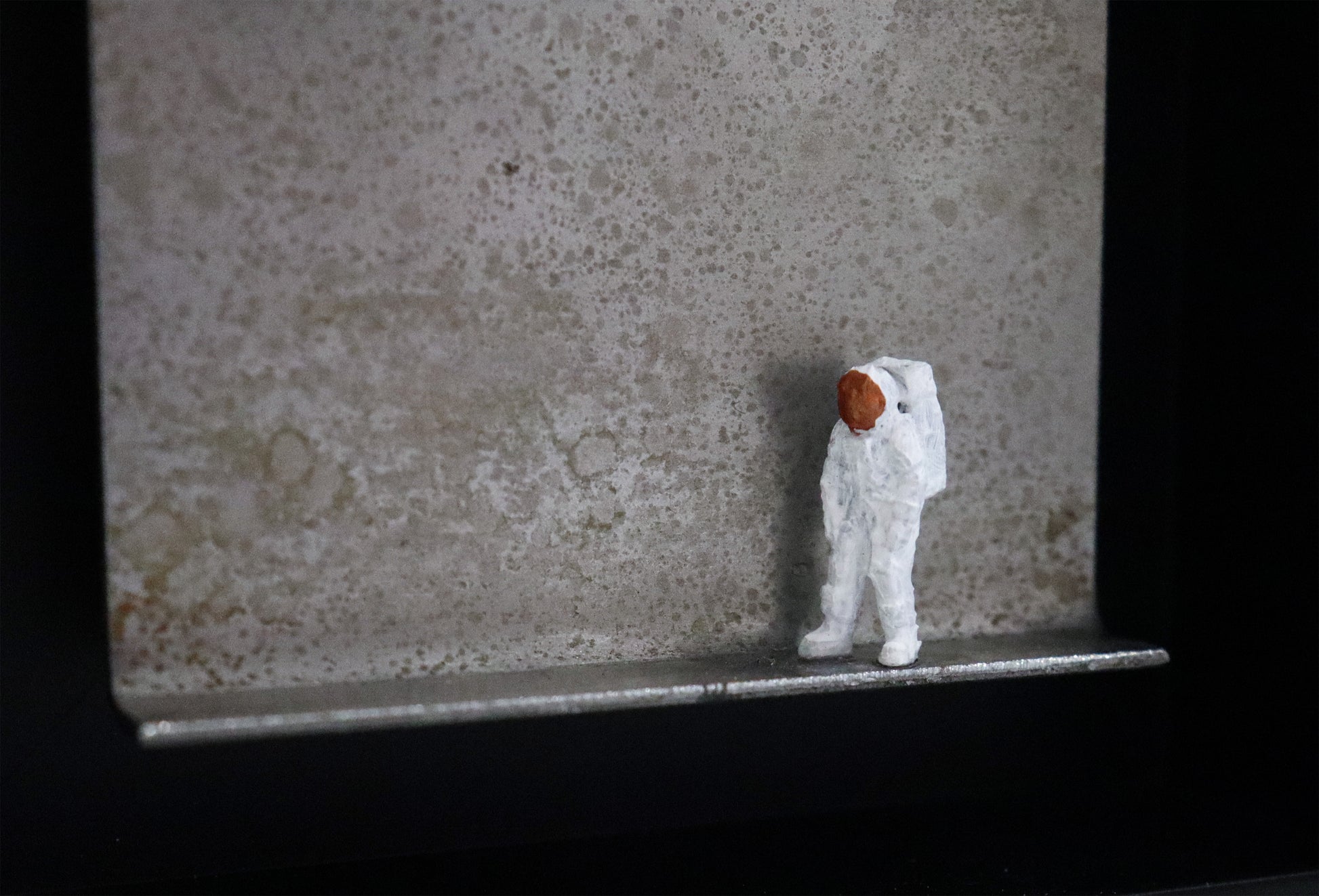 Original Objekt - Assemblage von Katrin Schramm, Titel „That's one  small step...“ – Metallwinkel mit Miniatur-Figur, Detailansicht. Dahinter das Metall des Winkels mit Struktur, als Hintergrund. Objektcollage im schwarzen Rahmen, erhältlich bei Galerie Waldbach - What The Artist Made