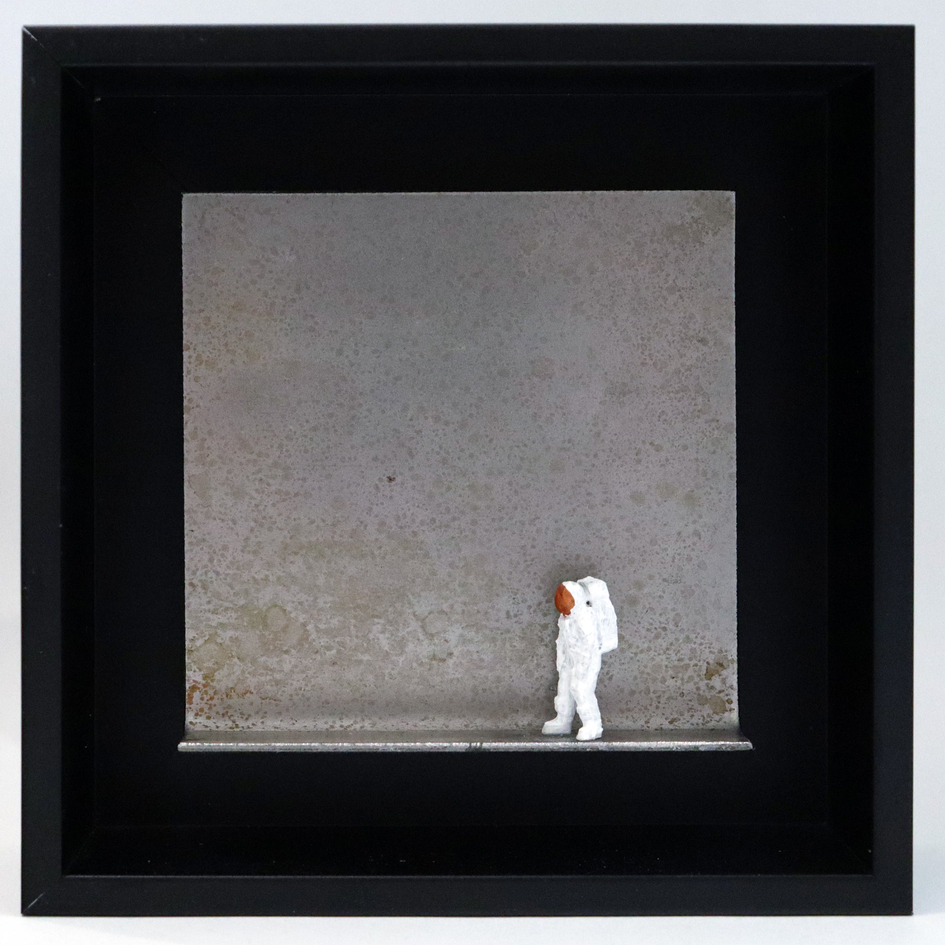 Original Objekt - Assemblage von Katrin Schramm, Titel „That's one  small step...“ – Metallwinkel mit Miniatur-Figur: Ein Astronaut steht am rechten Rand des Winkels und schaut zur gegenüberliegenden Seite. Dahinter das Metall des Winkels mit Struktur, als Hintergrund. Objektcollage im schwarzen Rahmen, erhältlich bei Galerie Waldbach - What The Artist Made