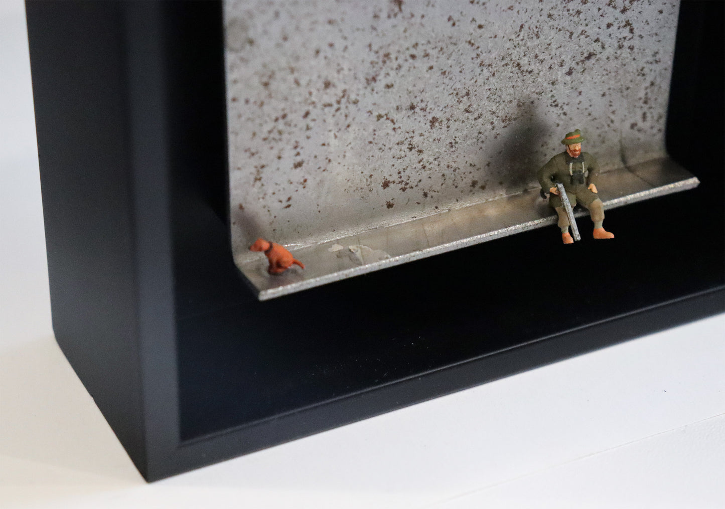 Original Objekt - Assemblage von Katrin Schramm, Titel „Waidmanslust“ – Metallwinkel mit Miniatur-Figuren, Detailansicht: Ein Jäger sitzt in Jägerkleidung an der Kante des Winkels und hält sein Gewehr in der Hand. Am linken Rand sitzt ein Hund und schaut aus dem Bildbereich. Dahinter das Metall des Winkels mit Struktur, als Hintergrund. In einem schwarzen Rahmen aus Holz. Objektcollage, erhältlich bei Galerie Waldbach - What The Artist Made