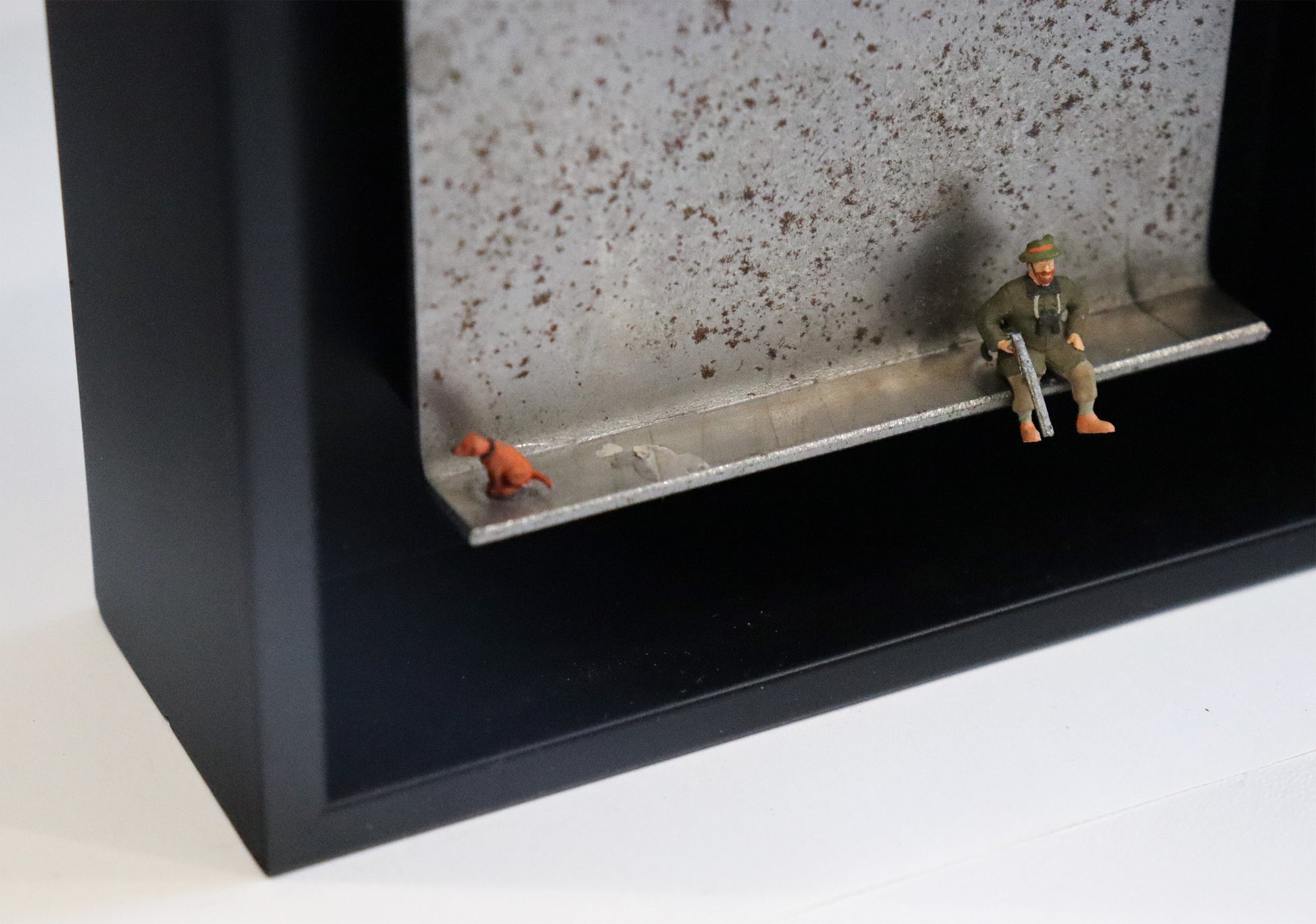 Original Objekt - Assemblage von Katrin Schramm, Titel „Waidmanslust“ – Metallwinkel mit Miniatur-Figuren, Detailansicht: Ein Jäger sitzt in Jägerkleidung an der Kante des Winkels und hält sein Gewehr in der Hand. Am linken Rand sitzt ein Hund und schaut aus dem Bildbereich. Dahinter das Metall des Winkels mit Struktur, als Hintergrund. In einem schwarzen Rahmen aus Holz. Objektcollage, erhältlich bei Galerie Waldbach - What The Artist Made