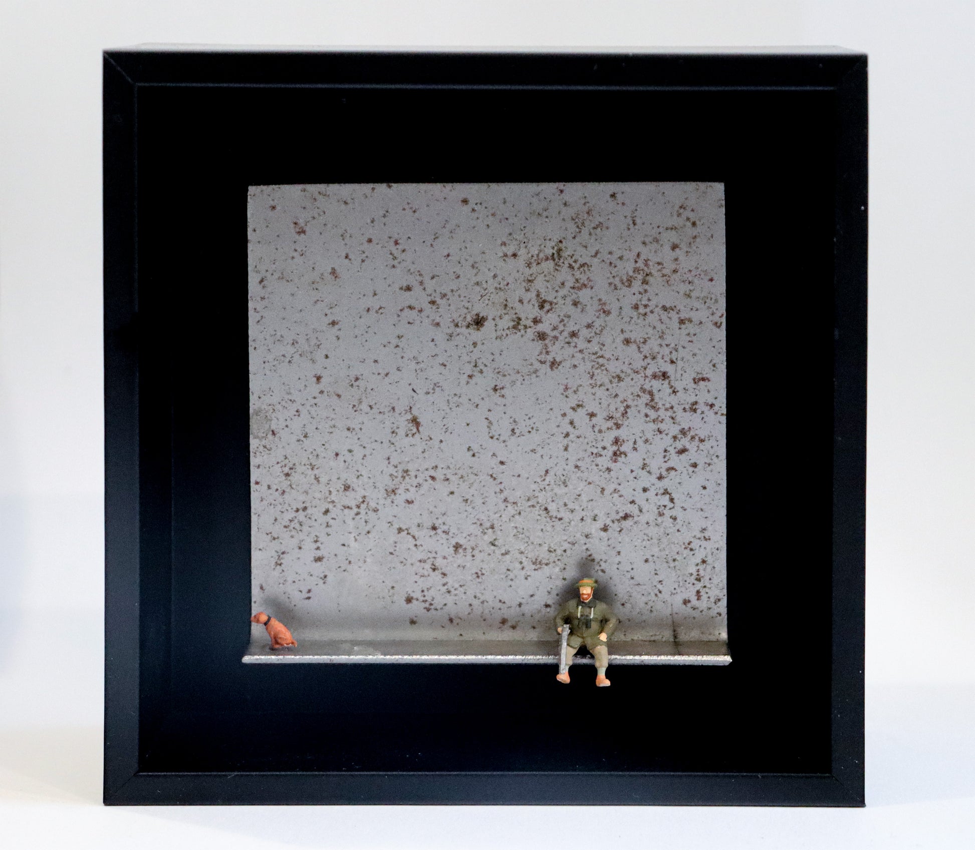 Original Objekt - Assemblage von Katrin Schramm, Titel „Waidmanslust“ – Metallwinkel mit Miniatur-Figuren: Ein Jäger sitzt in Jägerkleidung an der Kante des Winkels und hält sein Gewehr in der Hand. Am linken Rand sitzt ein Hund und schaut aus dem Bildbereich. Dahinter das Metall des Winkels mit Struktur, als Hintergrund. In einem schwarzen Rahmen aus Holz. Objektcollage, erhältlich bei Galerie Waldbach - What The Artist Made