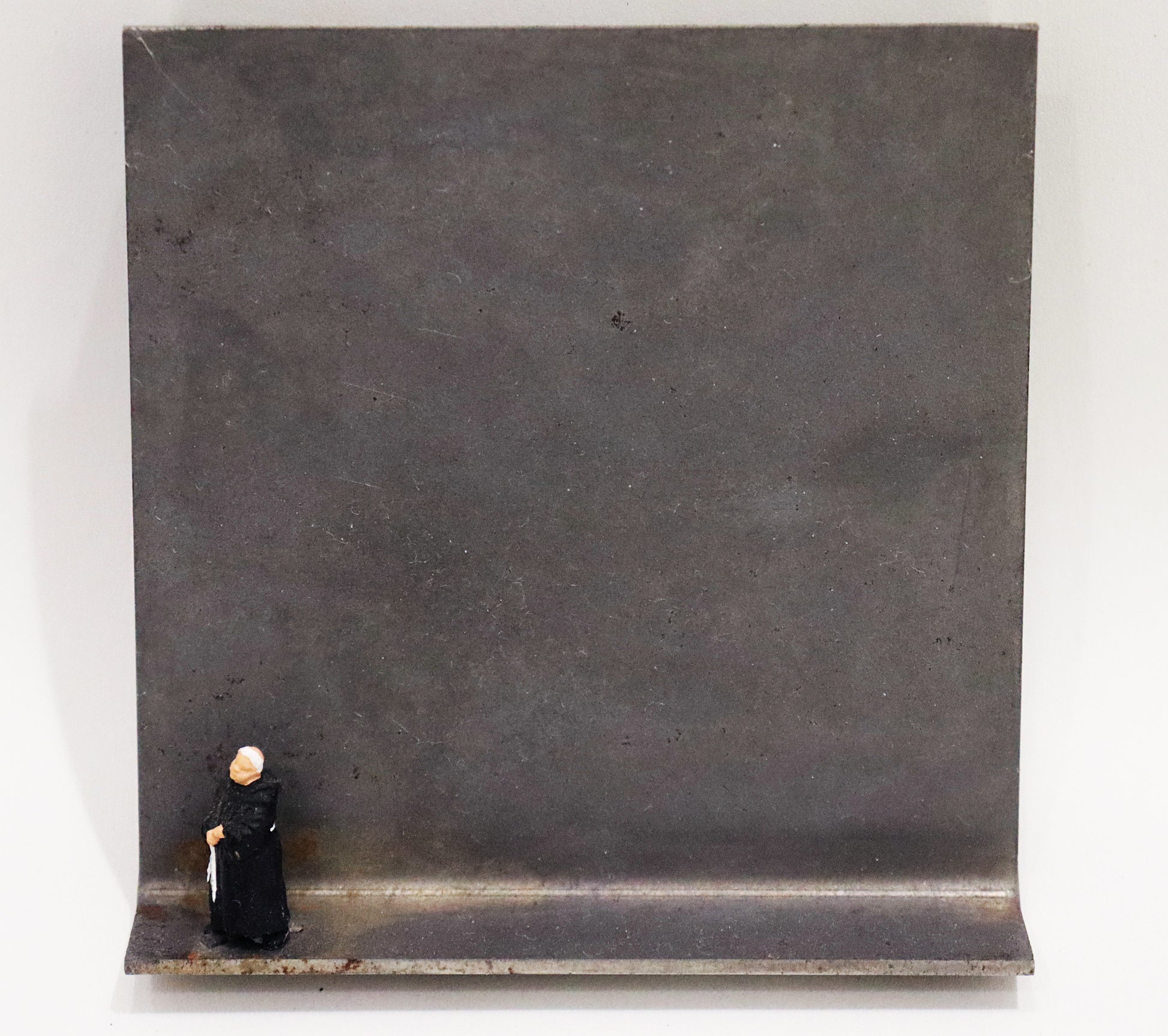 Original Objekt - Assemblage von Katrin Schramm, Titel „Wenn ich stehe, dann stehe ich.“ – Metallwinkel mit Miniatur-Figur: am linken Bildrand Mönch in schwarzem Habit stehend, mit gefalteten Händen und zur Seite schauend. Dahinter das Metall des Winkels mit Struktur, als Hintergrund. - Objektcollage, erhältlich bei Galerie Waldbach - What The Artist Made