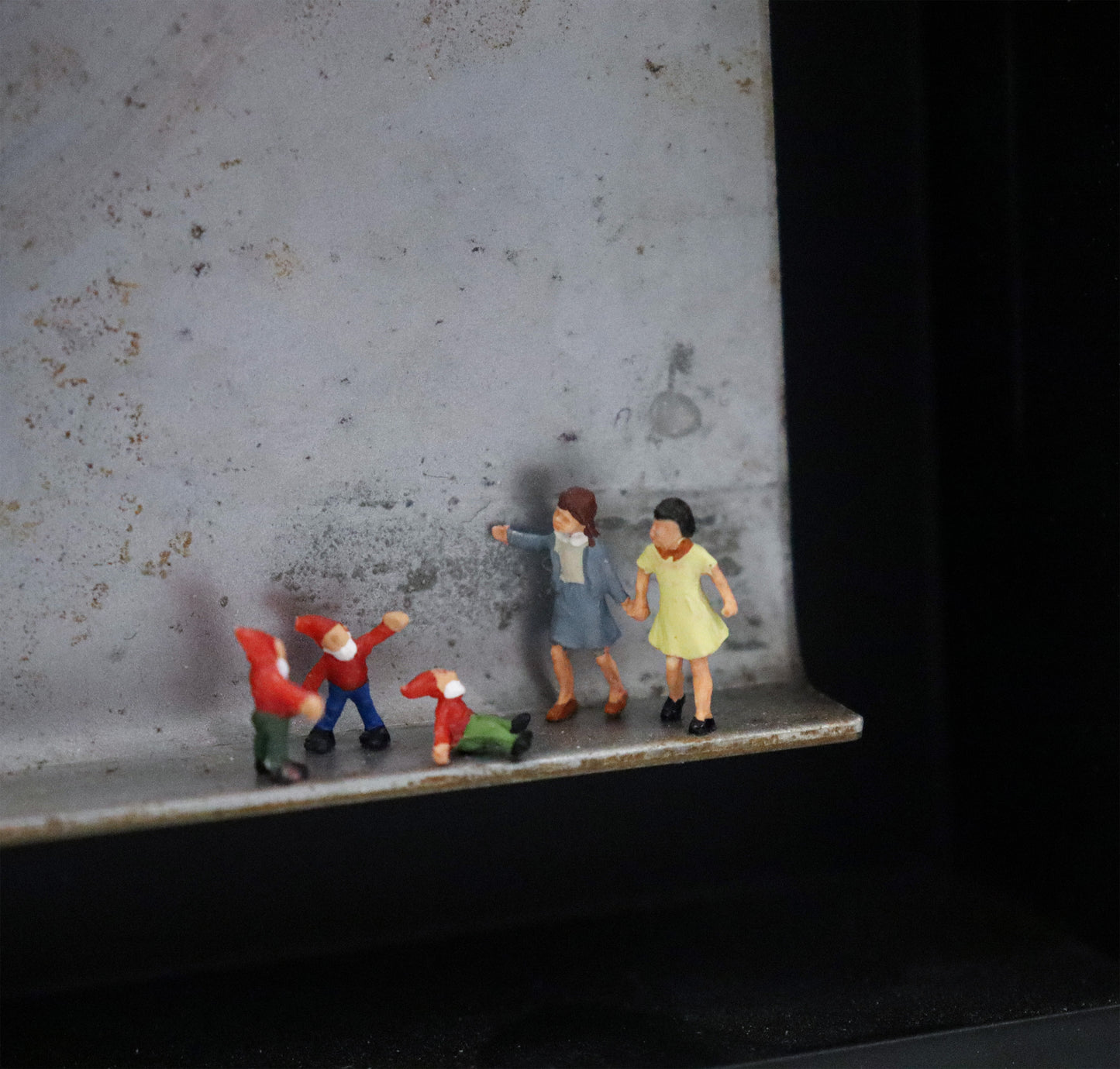 Original Objekt - Assemblage von Katrin Schramm, Titel „Zwergengeflüster.“ – Metallwinkel mit Miniatur-Figuren, Detailansicht der Gruppe. Dahinter das Metall des Winkels mit Struktur, als Hintergrund. Objektcollage im schwarzen Rahmen, erhältlich bei Galerie Waldbach - What The Artist Made