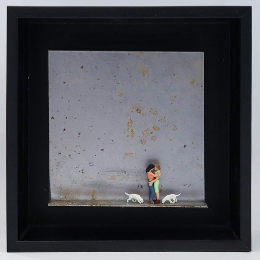 Original Objekt - Assemblage von Katrin Schramm, Titel „Bonding.“ – Metallwinkel mit Miniatur-Figuren: Zwei Frauen stehen einander in den Armen haltend auf dem Winkel. Links und rechts von den beiden stehen zwei weiße Hunde, die Köpfe suchend gesenkt. Dahinter das Metall des Winkels mit Struktur, als Hintergrund. Objektcollage im schwarzen Rahmen, erhältlich bei Galerie Waldbach - What The Artist Made