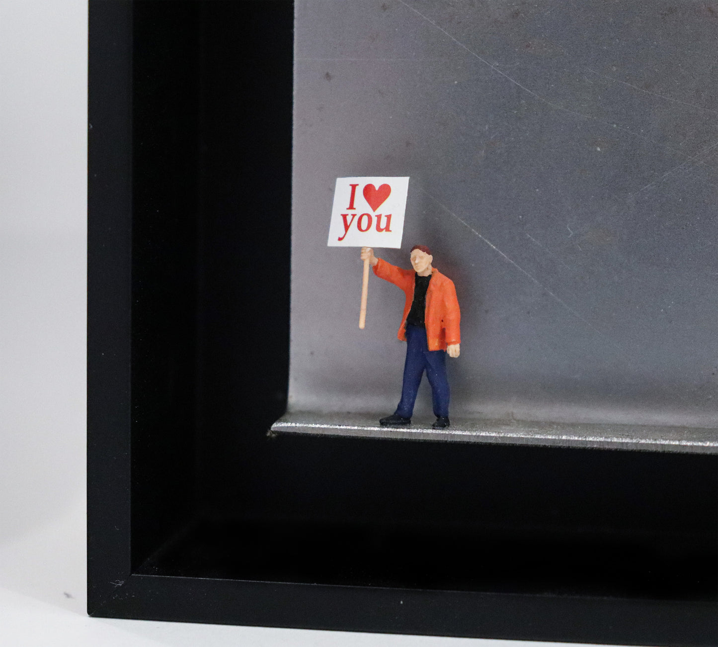 Original Objekt - Assemblage von Katrin Schramm, Titel „Einfach so!“ – Metallwinkel mit Miniatur-Figur, Detailansicht. Dahinter das Metall des Winkels mit Struktur, als Hintergrund. Objektcollage im schwarzen Rahmen, erhältlich bei Galerie Waldbach - What The Artist Made