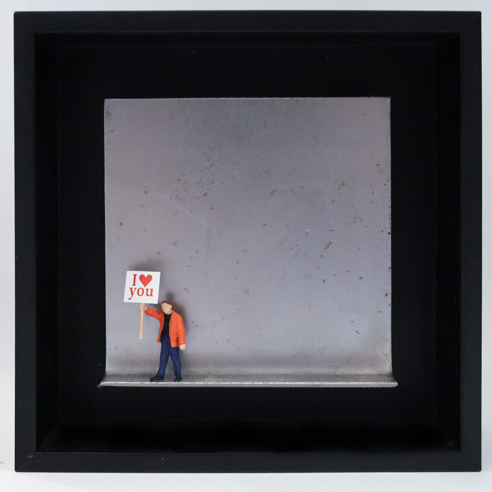 Original Objekt - Assemblage von Katrin Schramm, Titel „Einfach so!“ – Metallwinkel mit Miniatur-Figur: Eine Person in orangener Jacke steht am linken Rand des Winkels und hält ein Schild hoch. Darauf steht "I (heart) you". Dahinter das Metall des Winkels mit Struktur, als Hintergrund. Objektcollage im schwarzen Rahmen, erhältlich bei Galerie Waldbach - What The Artist Made