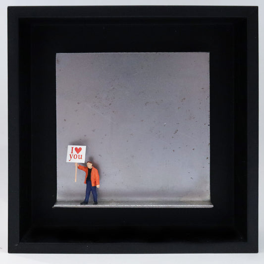 Original Objekt - Assemblage von Katrin Schramm, Titel „Einfach so!“ – Metallwinkel mit Miniatur-Figur: Eine Person in orangener Jacke steht am linken Rand des Winkels und hält ein Schild hoch. Darauf steht "I (heart) you". Dahinter das Metall des Winkels mit Struktur, als Hintergrund. Objektcollage im schwarzen Rahmen, erhältlich bei Galerie Waldbach - What The Artist Made