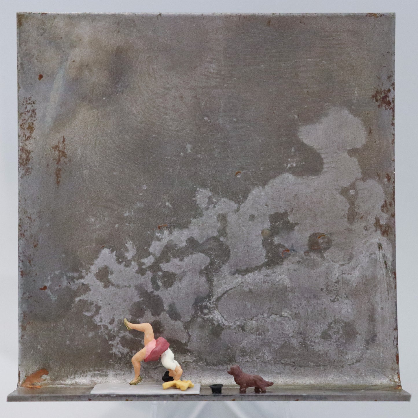 Original Objekt - Assemblage von Katrin Schramm, Titel „Weil sie s kann.“ – Metallwinkel mit Miniatur-Figuren: Eine Frau macht eine Brücke auf einer Yogamatte. Vor ihr ein handtuch und ein Hut, sowie ein kleiner brauner Hund, der sie anschaut. Dahinter das Metall des Winkels mit Struktur, als Hintergrund. Objektcollage, erhältlich bei Galerie Waldbach - What The Artist Made