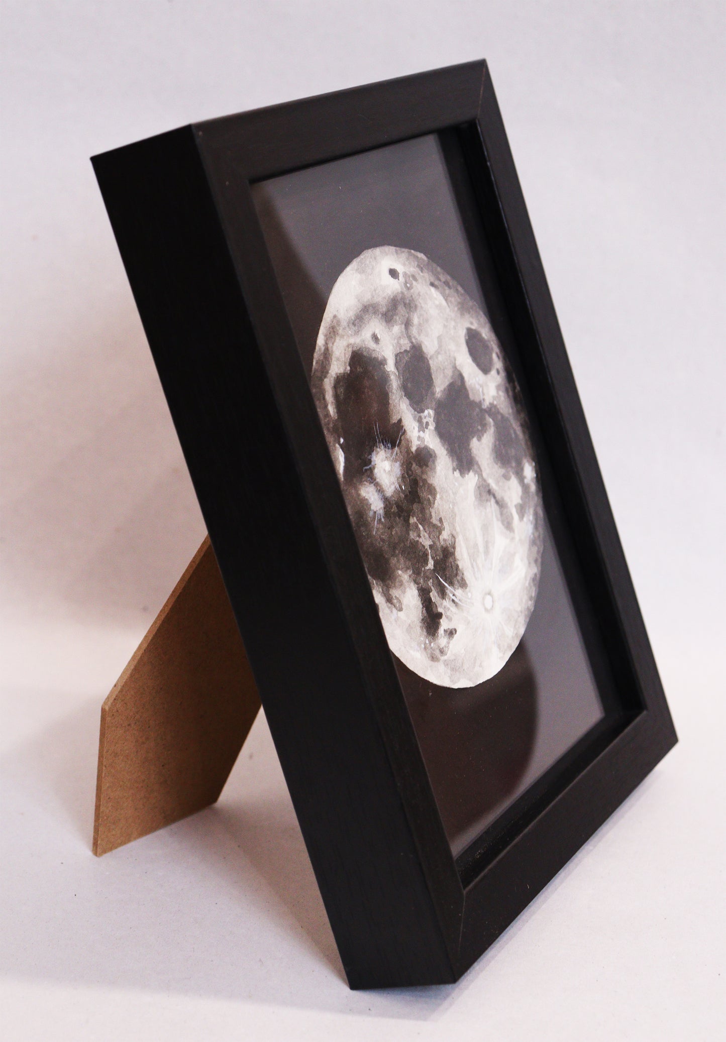 Original Aquarell-Malerei, Gemälde von Lisa  Steinbrück, Titel „Luna“ – Darstellung des Mondes der Erde in Aquarell-Stil, schwarz-weiß und Grautöne auf schwarzem Grund, Aquarell auf Papier, gerahmt, schwarzer Rahmen, Seitenansicht auf den stehenden Rahmen - erhältlich bei Galerie Waldbach - What The Artist Made
