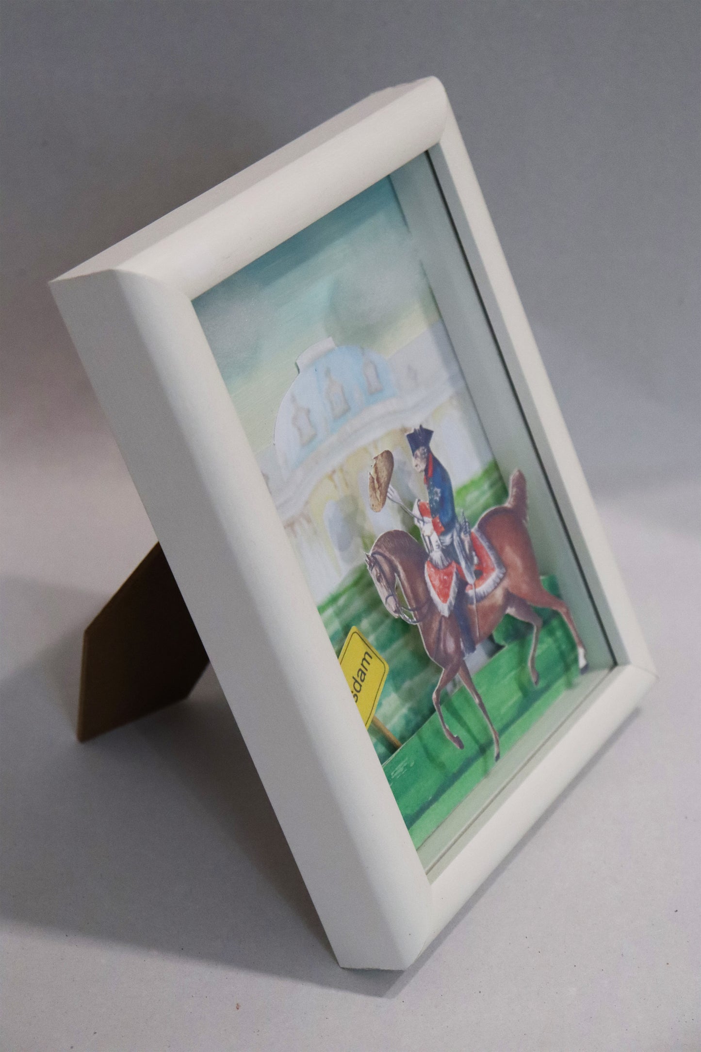Collage  nach einer Malerei von Nadine Conrad, Titel „Kartoffelfritze“ – Friedrich II auf Pferd reitend, er hält eine große Gabel wie eine Lanze, darauf steckt eine große Kartoffel. Links von ihm ist das Ortsschild von Potsdam, im Hintergrund das Schloss Sanssouci, Papiercollage, gerahmt -  Seitenansicht auf den stehenden weißen Rahmen.   Erhältlich bei Galerie Waldbach - What The Artist Made
